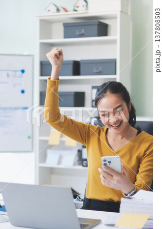 Asian woman works on laptop at office desk. She smiles happily while typing on keyboard near window with blinds. Professional woman works in business environment. Asian woman works on laptop at office desk. She smiles happily while typing on keyboard near window with blinds. Professional woman works in business environment. 137808503