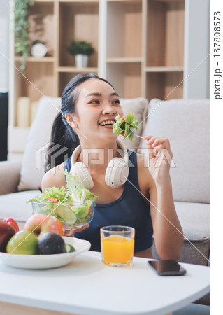 Fit young Asian women in activewear sitting on yoga mat and eating fresh vegetable salad after workout, representing clean eating, fitness lifestyle, wellness routine and health balance. 137808573
