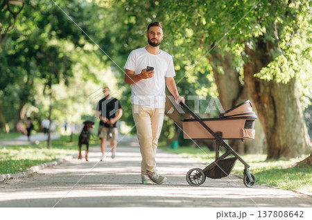 Smartphone in hand. Father is with cradle, having a walk in the park Smartphone in hand. Father is with cradle, having a walk in the park 137808642