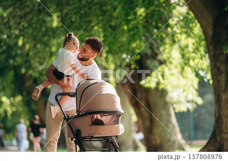 Girl is in hand of dad. Man with his daughter and cradle in the park Girl is in hand of dad. Man with his daughter and cradle in the park 137808976
