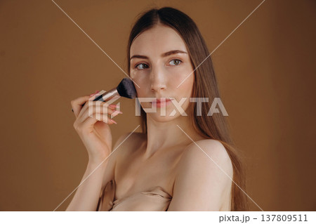 Using brush, cosmetics on face. Young woman is against background Using brush, cosmetics on face. Young woman is against background 137809511