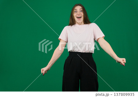Various emotions of a European woman in a pink T-shirt and black pants on a green background with long brown hair in different poses 137809935