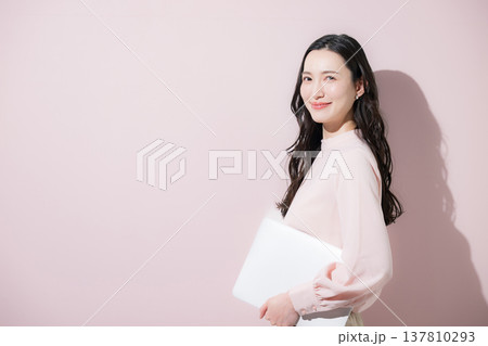 ピンクの背景でパソコンを持つかわいいオフィスカジュアルの日本人女性笑顔のカメラ目線 137810293