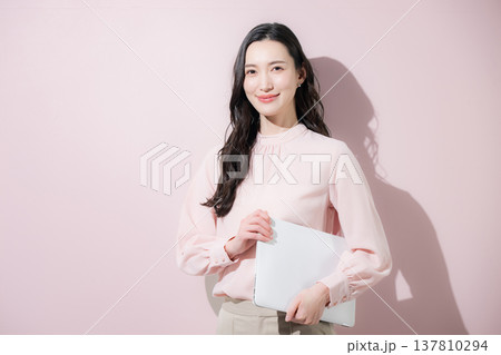 ピンクの背景でパソコンを持つかわいいオフィスカジュアルの日本人女性笑顔のカメラ目線 137810294