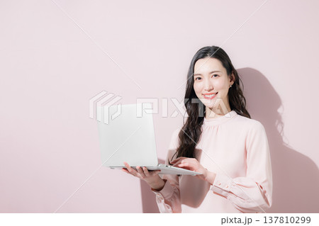 ピンクの背景でパソコンを持つオフィスカジュアルの日本人女性笑顔のカメラ目線 2026トレンドカラー ピンクの背景でパソコンを持つオフィスカジュアルの日本人女性笑顔のカメラ目線 2026トレンドカラー 137810299