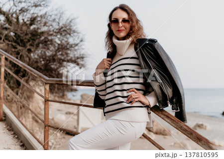 Woman fashion beach stylish model in striped sweater and leather jacket posing by a railing at the coast during autumn Woman fashion beach stylish model in striped sweater and leather jacket posing by a railing at the coast during autumn 137810595