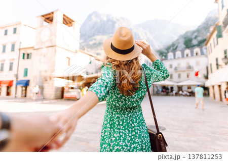 Young woman with curly hair playfully adjusts her wide-brimmed hat while strolling through a square 137811253