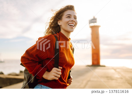 Young woman enjoying a breezy day near a vibrant red lighthouse. Autumn sunset 137811616