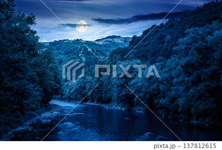 river flowing through green forest at the foot of mountain scenery at night. full moon over mysterious landscape of countryside in summer. nature picture with stones on shore and cloudy sky 137812615