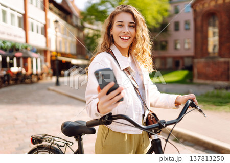 Woman using mobile phone, walks with bicycle, looks around at beautiful architecture. Selfie time. 137813250