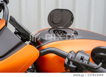 A horizontal shot from a side perspective showing the open charging socket on an orange electric motorcycle, with the handlebars and digital instrument cluster in the foreground against a neutral back A horizontal shot from a side perspective showing the open charging socket on an orange electric motorcycle, with the handlebars and digital instrument cluster in the foreground against a neutral back 137813444