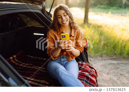 Young curly woman in stylish clothes in the trunk of a car in the forest and uses the phone. 137813625