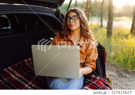 Happy woman working with laptop from the car trunk or boot in camper sunny summer park. Happy woman working with laptop from the car trunk or boot in camper sunny summer park. 137813627