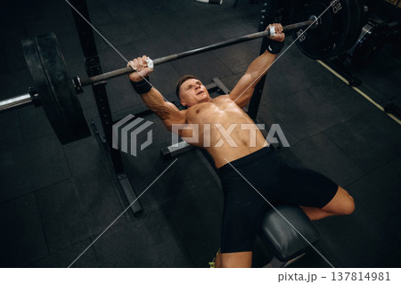 Pushing the barbell, lying down. Athletic man performing weightlifting exercises in a gym Pushing the barbell, lying down. Athletic man performing weightlifting exercises in a gym 137814981