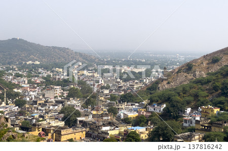 Jaipur cityscape fills valley beneath fortified hill and pale sky. Historic Rajasthan fort stands 137816242