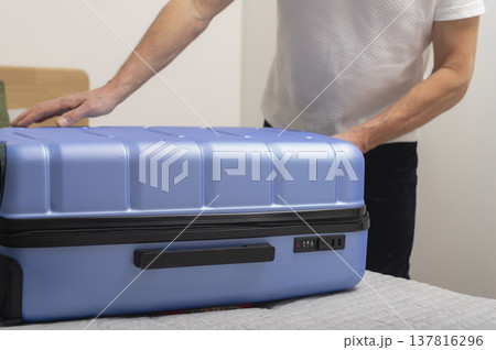 Man pressing blue suitcase lid to close luggage. Male securing hardcase baggage at home before trip. Travel organization, packing completion, departure readiness concept.. Man pressing blue suitcase lid to close luggage. Male securing hardcase baggage at home before trip. Travel organization, packing completion, departure readiness concept.. 137816296