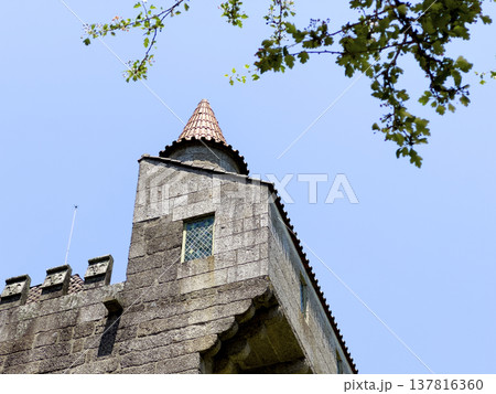 Stone castle tower with tiled roof and small window in Guimaraes Portugal. Medieval architecture rising against clear blue sky framed by green branches. Travel destination, historic heritage 137816360