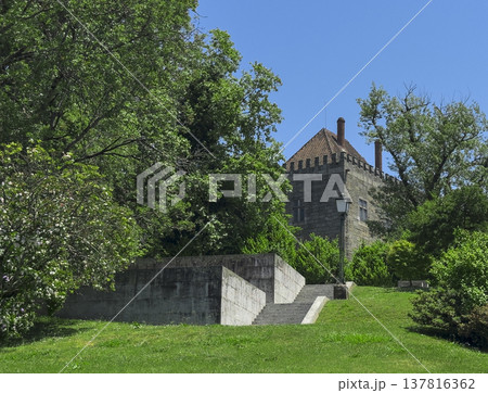 Stone medieval tower surrounded by green trees in Guimaraes Portugal. Historic fortress with tiled roof rising above park lawn under blue sky. Travel destination, European heritage, scenic 137816362