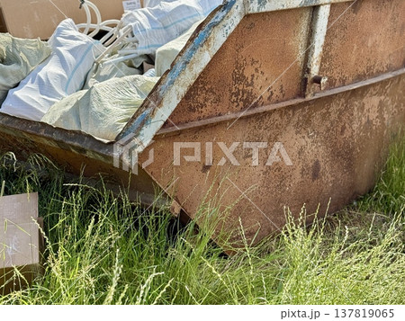 An Old and Rusty Dump Container That is Surrounded by Lush, Overgrown Grass and Weeds. A weathered metal dumpster overflowing with various debris, positioned against a backdrop of lush tall grass. An Old and Rusty Dump Container That is Surrounded by Lush, Overgrown Grass and Weeds. A weathered metal dumpster overflowing with various debris, positioned against a backdrop of lush tall grass. 137819065