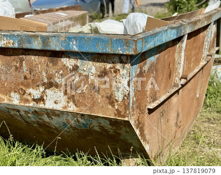 An Old and Rusty Dump Container That is Surrounded by Lush, Overgrown Grass and Weeds. A weathered metal dumpster overflowing with various debris, positioned against a backdrop of lush tall grass. 137819079
