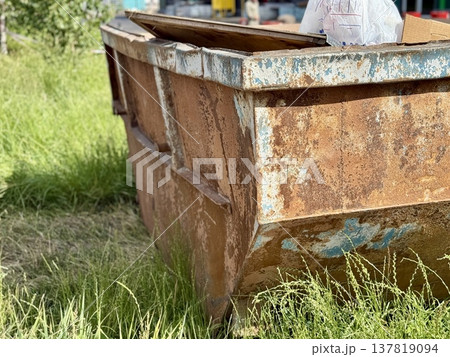 An Old and Rusty Dump Container That is Surrounded by Lush, Overgrown Grass and Weeds. A weathered metal dumpster overflowing with various debris, positioned against a backdrop of lush tall grass. 137819094