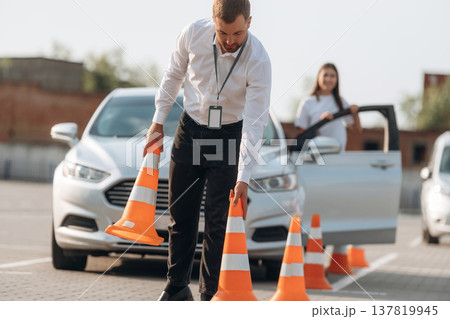 Placing the guide cones. Man instructor in the driving school and woman are outside the car Placing the guide cones. Man instructor in the driving school and woman are outside the car 137819945