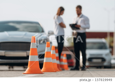 Talking with each other, cones on the road. Man instructor in the driving school and woman are outside the car Talking with each other, cones on the road. Man instructor in the driving school and woman are outside the car 137819949