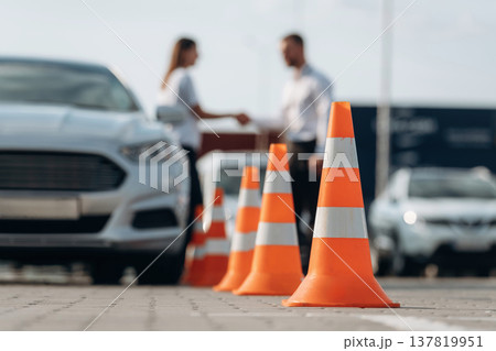 Talking with each other, cones on the road. Man instructor in the driving school and woman are outside the car Talking with each other, cones on the road. Man instructor in the driving school and woman are outside the car 137819951