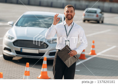 Notepad is in hand. Driving school worker is outdoors near car Notepad is in hand. Driving school worker is outdoors near car 137820181