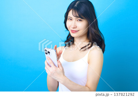 スマートフォンを持ち笑顔でこちらを見つめる若い日本人女性 青背景 137821292