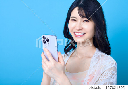 スマートフォンを持って笑顔を見せる若い日本人女性 青背景 コピースペース スマートフォンを持って笑顔を見せる若い日本人女性 青背景 コピースペース 137821295