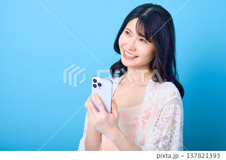 青い背景でスマートフォンを持ち笑顔で未来をイメージする日本人女性 青い背景でスマートフォンを持ち笑顔で未来をイメージする日本人女性 137821393