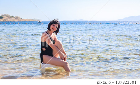 Elegant woman in a black swimsuit sitting on a plastic chair, enjoying the serene sea view, capturing the essence of summer relaxation and beach vibes 137822301
