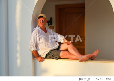 Adult man in a white shirt and cap sitting on a wide windowsill, enjoying the beautiful sunset, reflecting on life and tranquility in the golden hour Adult man in a white shirt and cap sitting on a wide windowsill, enjoying the beautiful sunset, reflecting on life and tranquility in the golden hour 137822368