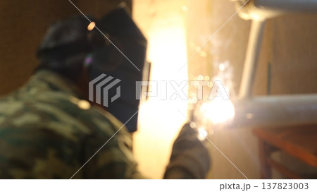 A welder, seen from the back, wearing a uniform and protective helmet, welds a house heating radiator made from white-painted iron pipes, a blurred image of an unrecognizable welder in the process 137823003