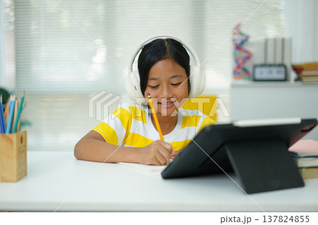 Asian Girl with Headphones Taking Notes While Watching Online Class on Tablet Asian Girl with Headphones Taking Notes While Watching Online Class on Tablet 137824855