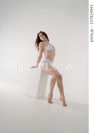 Graceful young woman in white lingerie posing on a cube in a minimalist high-key studio. Graceful young woman in white lingerie posing on a cube in a minimalist high-key studio. 137824943