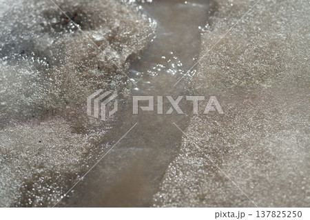 Spring flow over icy slush. Brown liquid and coarse gravel flow through melting icy slush. Muddy streams with gritty water trickle over melting ice in urban setting Spring flow over icy slush. Brown liquid and coarse gravel flow through melting icy slush. Muddy streams with gritty water trickle over melting ice in urban setting 137825250