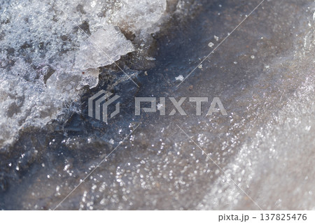 Translucent ice sheets disintegrate into fragments beside flowing river. Delicate layers of ice fracture into small pieces along quiet streambank during spring thaw 137825476