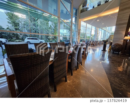 A luxurious hotel restaurant interior featuring elegant wicker chairs, neatly set tables with red napkins, and large floor-to-ceiling glass windows overlooking a lush green tropical garden view. A luxurious hotel restaurant interior featuring elegant wicker chairs, neatly set tables with red napkins, and large floor-to-ceiling glass windows overlooking a lush green tropical garden view. 137826419