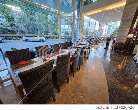 A luxurious hotel restaurant interior featuring elegant wicker chairs, neatly set tables with red napkins, and large floor-to-ceiling glass windows overlooking a lush green tropical garden view. 137826420