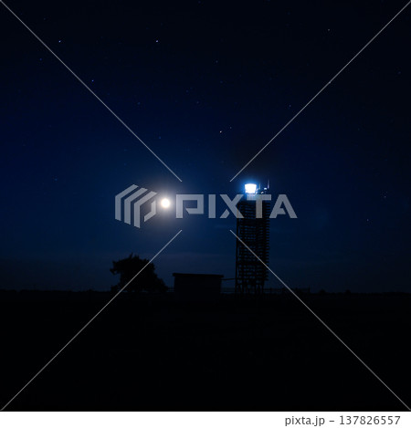 Silhouette of a navigation tower and a lonely tree against a deep blue night sky with stars and a bright moon. Peaceful maritime concept of safety, guidance, and hope in the dark. Silhouette of a navigation tower and a lonely tree against a deep blue night sky with stars and a bright moon. Peaceful maritime concept of safety, guidance, and hope in the dark. 137826557