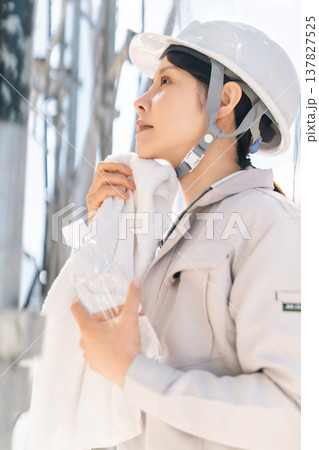 夏の建設現場での熱中症対策。作業の合間にタオルで汗を拭い水分補給をする女性職人 137827525