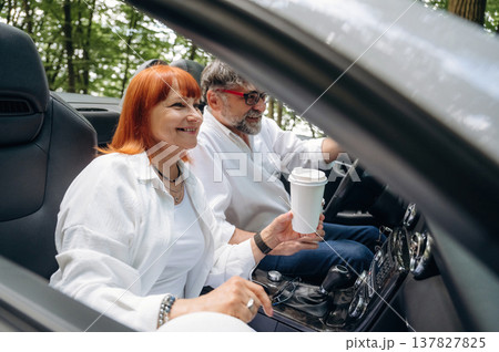 Cup of drink in woman's hand. Senior couple driving convertible cabriolet car Cup of drink in woman's hand. Senior couple driving convertible cabriolet car 137827825