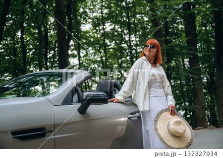 Woman with hat in hand is standing near cabriolet car on the road against trees 137827934