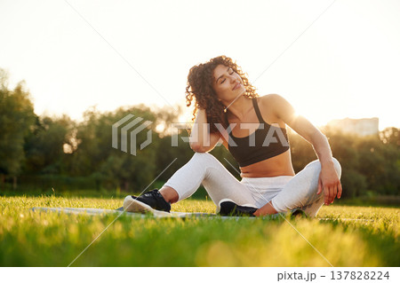 Just resting, sitting on yoga mat and smiling. Young fitness woman is on the field outdoors Just resting, sitting on yoga mat and smiling. Young fitness woman is on the field outdoors 137828224