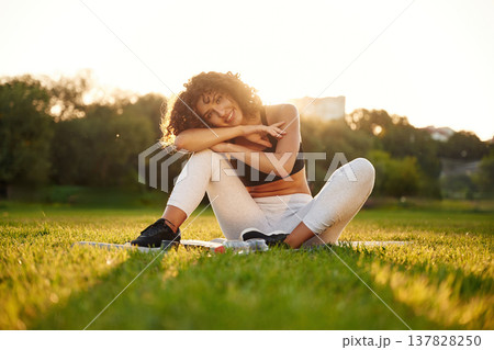 Just resting, sitting on yoga mat and smiling. Young fitness woman is on the field outdoors Just resting, sitting on yoga mat and smiling. Young fitness woman is on the field outdoors 137828250