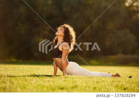 In black top and white pants, doing back stretching. Young fitness woman is on the field outdoors In black top and white pants, doing back stretching. Young fitness woman is on the field outdoors 137828284