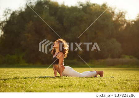 In black top and white pants, doing back stretching. Young fitness woman is on the field outdoors In black top and white pants, doing back stretching. Young fitness woman is on the field outdoors 137828286
