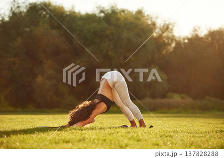 In black top and white pants, doing back stretching. Young fitness woman is on the field outdoors In black top and white pants, doing back stretching. Young fitness woman is on the field outdoors 137828288